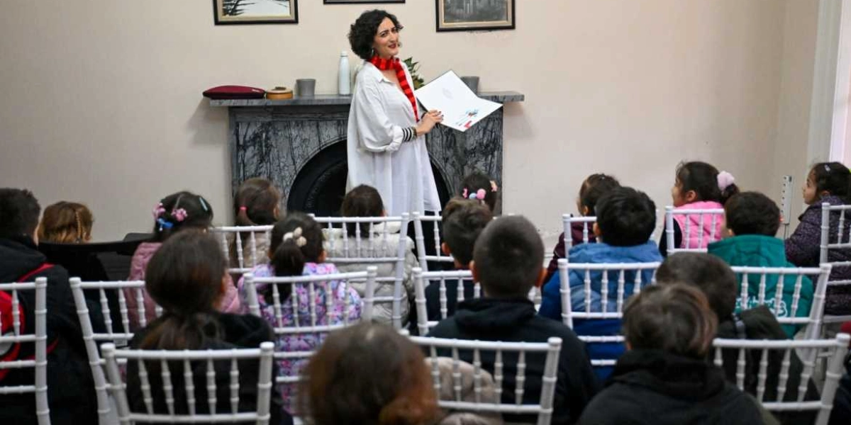 Kıvırcık saçlı, beyaz gömlekli ve kırmızı çizgili atkılı bir kadın, mermer bir şöminenin önünde durarak çocuklara kitap okuyor. Çocuklar, beyaz sandalyelerde oturmuş, ilgiyle kadını dinliyor.
