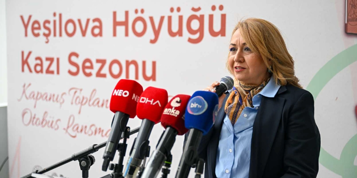 Yeşilova Höyüğü Kazı Sezonu Kapanış Toplantısı ve Otobüs Lansmanı etkinliğinde, kürsüde konuşma yapan sarı saçlı bir kadın, önünde sıralanmış farklı basın kuruluşlarına ait mikrofonlara hitap ediyor. Kadın, lacivert bir ceket ve açık mavi bir gömlek giymiş, boynunda desenli bir fular takıyor. Arka planda üzerinde etkinliğin adının yazılı olduğu beyaz bir pano bulunuyor.
