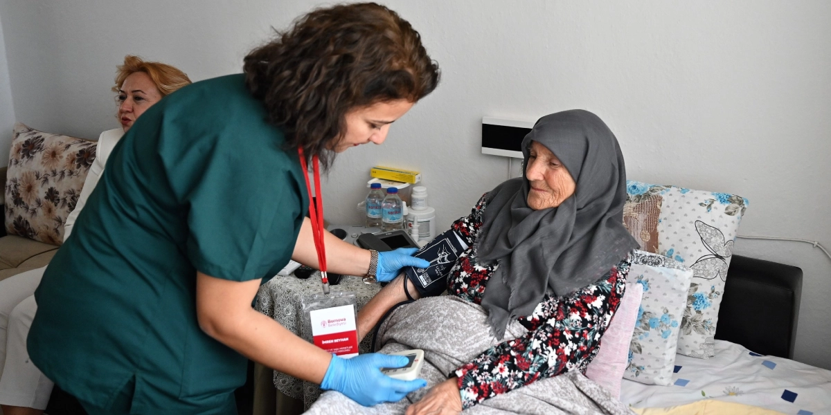 Yeşil üniformalı bir sağlık çalışanı, yatakta istirahat eden yaşlı bir kadının tansiyonunu ölçüyor. Sağlık çalışanı, üzerinde Bornova Belediyesi kimlik kartı asılı olan koyu yeşil bir forma giyiyor ve mavi eldivenlerle hastanın koluna tansiyon aleti manşonu takıyor. Başörtülü ve çiçekli bir bluz giyen yaşlı kadın, yatakta oturur vaziyette sağlık çalışanına bakıyor. Arka planda ise koltukta oturan başka bir kadın daha bulunuyor.