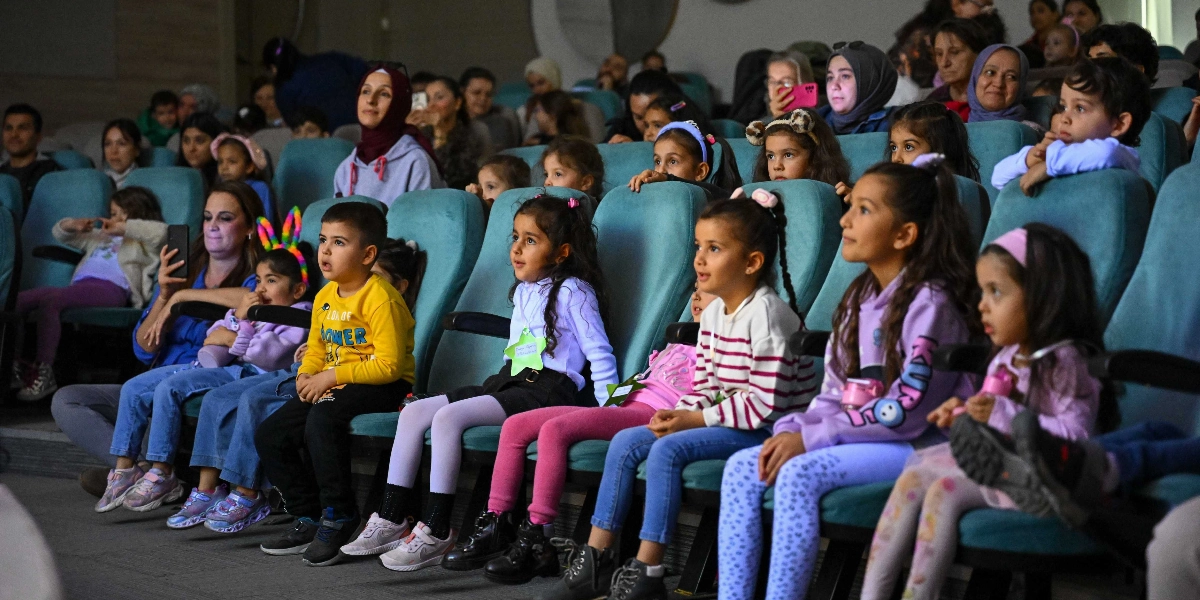Bir tiyatro veya konferans salonunda, turkuaz renkli koltuklarda yan yana oturmuş, ilgiyle sahneye bakan çocuklar ve onlara eşlik eden yetişkinler. Ön planda sarı tişörtlü bir erkek çocuk, beyaz bluzlu ve pembe pantolonlu bir kız çocuğu ile mor sweatshirt giyen başka bir kız çocuğu dikkat çekiyor. Çocukların birçoğu meraklı ve heyecanlı ifadelerle karşıya bakarken, arkalarında oturan ebeveynler de etkinliği takip ediyor.