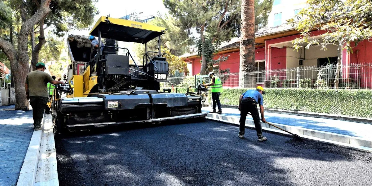 Sarı renkli bir asfalt serim makinesinin 159 Sokak'ta asfalt dökme işlemi yaptığı çalışma alanında, işçiler asfaltı düzeltmek için kürek ve tırmıklarla çalışıyor. Arka planda pembe renkli bir bina ve ağaçlar bulunuyor.