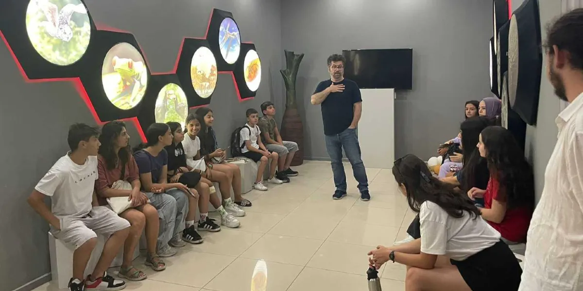 Modern bir müze veya bilim merkezinde, duvarda altıgen formlu ışıklı panellerin önünde oturan bir grup genç, ayakta duran bir eğitmeni dinliyor. Işıklı panellerde kurbağa, kuş ve böcek gibi doğa görselleri yer alıyor. Eğitmen, elini göğsüne koymuş bir şekilde gençlere hitap ederken, gençler dikkatle onu takip ediyor. Ortamda gri duvarlar, beyaz zemin ve dekoratif bir ağaç figürü bulunuyor.