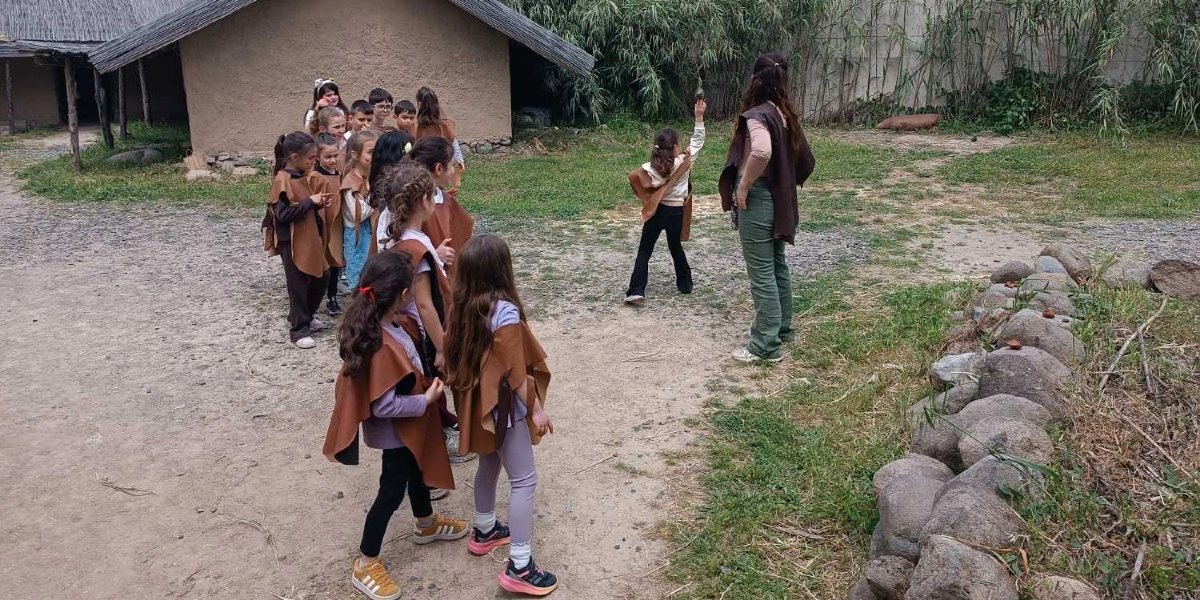Taş Devri temalı bir etkinlik alanında, üzerlerinde deri görünümlü pelerinler olan bir grup çocuk ve onlara eşlik eden bir yetişkin bulunuyor. Çocuklardan biri elindeki taşı havaya kaldırarak fırlatmaya hazırlanırken, diğerleri onu izliyor. Arka planda sazdan çatılı, kerpiç görünümlü tarih öncesi bir ev yapısı ve yeşil bitkiler yer alıyor.