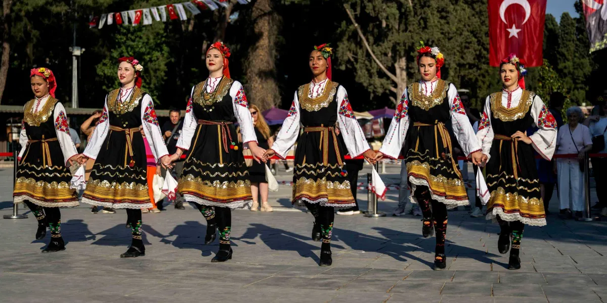 Geleneksel kıyafetler giymiş altı genç kadın, açık havada el ele tutuşarak halay çekiyor. Kadınlar, beyaz işlemeli bluzlar, siyah yelekler ve altın sarısı şeritlerle süslü siyah eteklerden oluşan yöresel kostümler giyiyor; saçlarında çiçekli taçlar bulunuyor. Arka planda Türk bayrağı ve ağaçların olduğu bir meydanda gerçekleşen gösteriyi izleyen insanlar görülüyor.