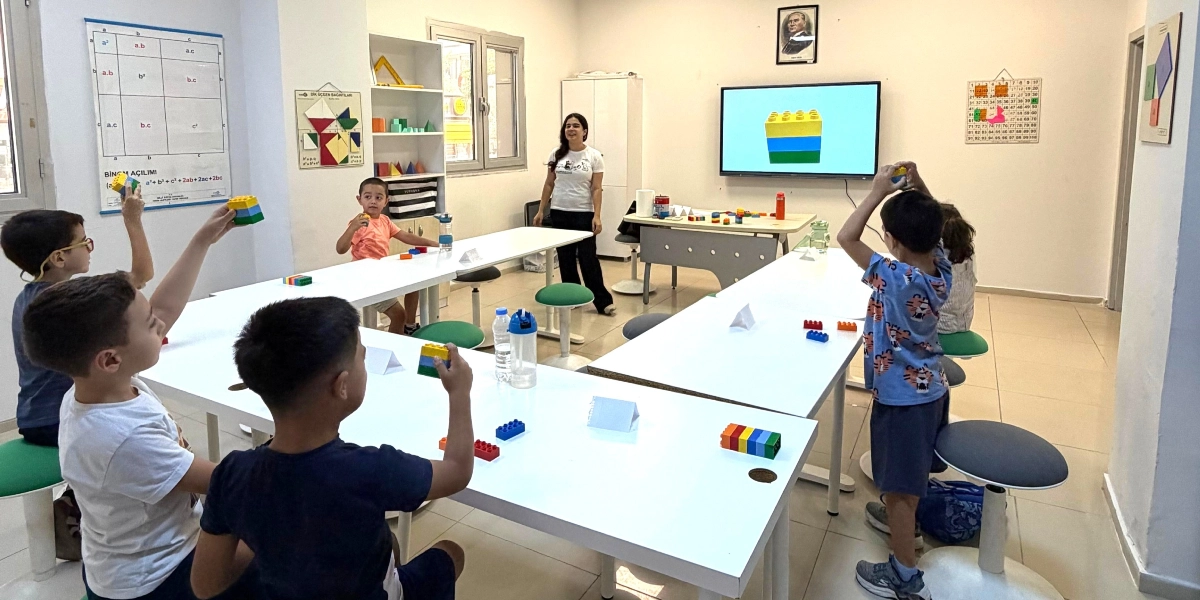 Aydınlık bir sınıfta, beyaz masaların etrafında oturan ve ayakta duran çocuklar, ellerindeki renkli yapı bloklarıyla çeşitli şekiller oluşturuyor. Sınıfın önünde duran bir eğitmen çocuklara rehberlik ederken, arkadaki akıllı tahtada da benzer bir yapı bloğu görseli yer alıyor. Duvarlarda matematiksel formüllerin ve geometrik şekillerin bulunduğu eğitim posterleri asılı.