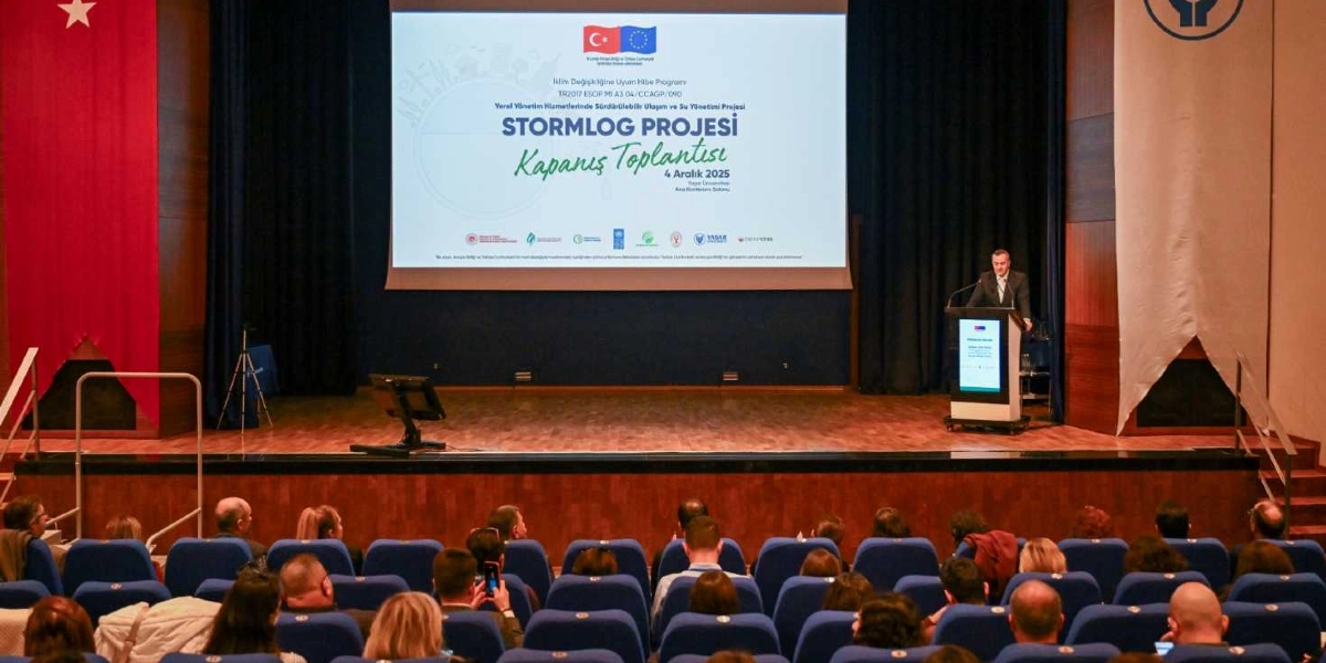 Konferans salonunda, sahnede yer alan kürsüde konuşma yapan bir kişi ve onu izleyen katılımcıların bulunduğu bir toplantı. Sahnenin arkasındaki dev ekranda 