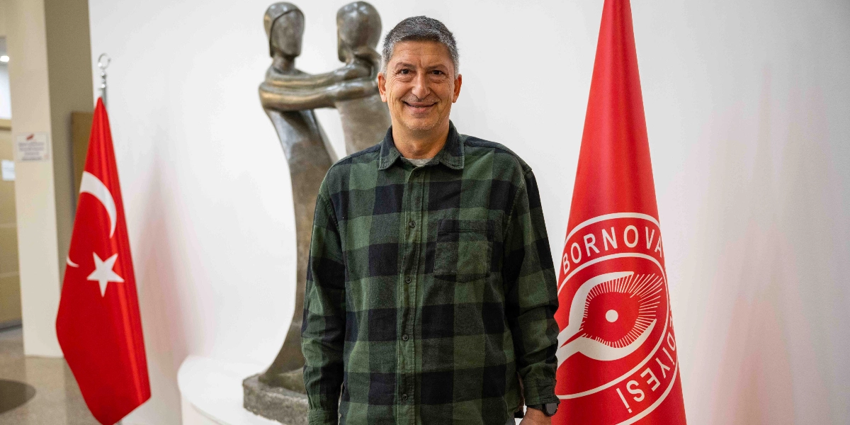 Yeşil ve siyah ekose desenli gömlek giymiş, gülümseyen bir erkek; arka planda sol tarafta Türk bayrağı, sağ tarafta üzerinde Bornova Belediyesi logosu bulunan kırmızı bir bayrak ve ortada birbirine sarılmış iki figürü temsil eden bronz renkli bir heykel ile poz veriyor.