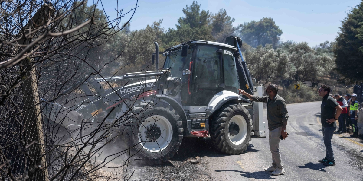 Bornova Homeros Vadisi'ndeki yangın bölgesinde, Hidromek marka bir iş makinesi yanmış çalılıkların olduğu alanda çalışma yaparken, yanındaki yolda duran iki kişi çalışmaları yönlendiriyor. İş makinesinin yanında duran kişi elindeki su şişesiyle alanı işaret ederken, yanındaki diğer kişi onu izliyor. Arka planda ise ağaçlık bir alan ve yol kenarında bekleyen görevliler görülüyor.