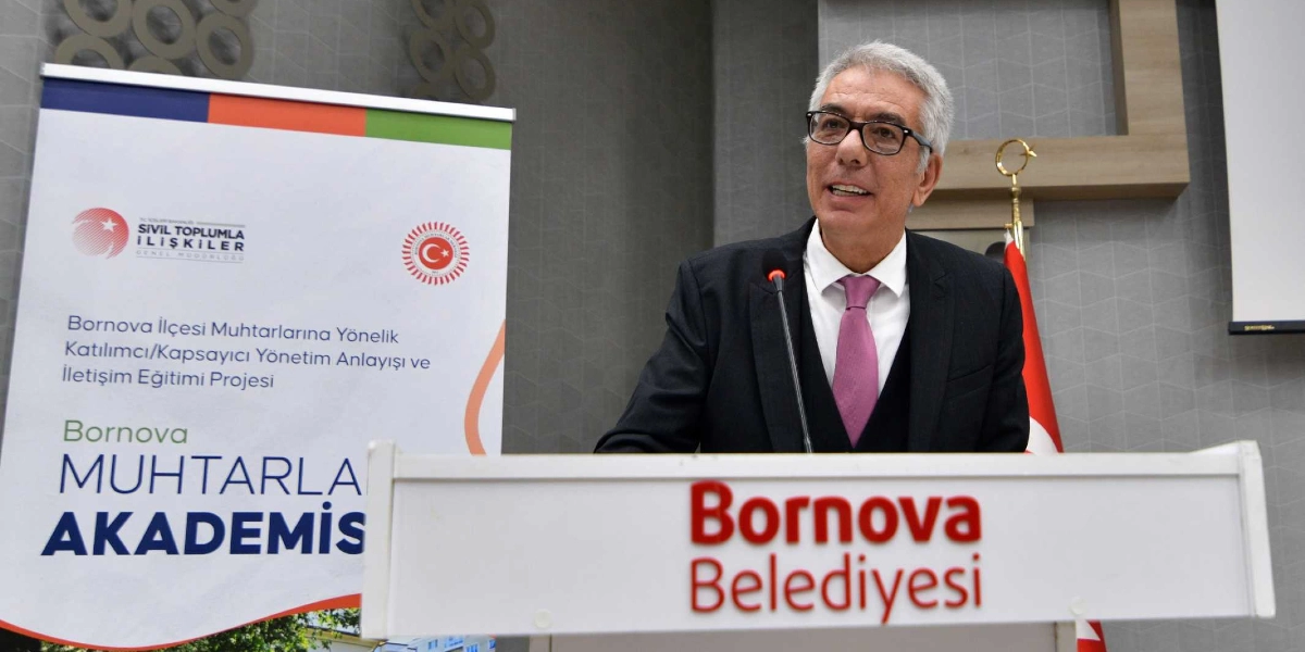 Kürsüde konuşma yapan, gözlüklü, gri saçlı ve takım elbiseli bir erkek. Kürsünün ön yüzünde kırmızı renkle 