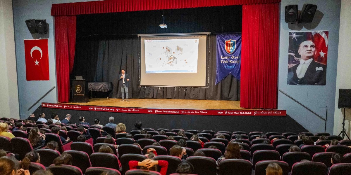 Konferans salonunda sahne üzerinde konuşma yapan bir kişi, arkasındaki dev ekranda deprem anında alınacak önlemleri gösteren bir sunum yapıyor. Sahnenin solunda Türk bayrağı, sağında ise Atatürk posteri asılı. Sahne kenarında 