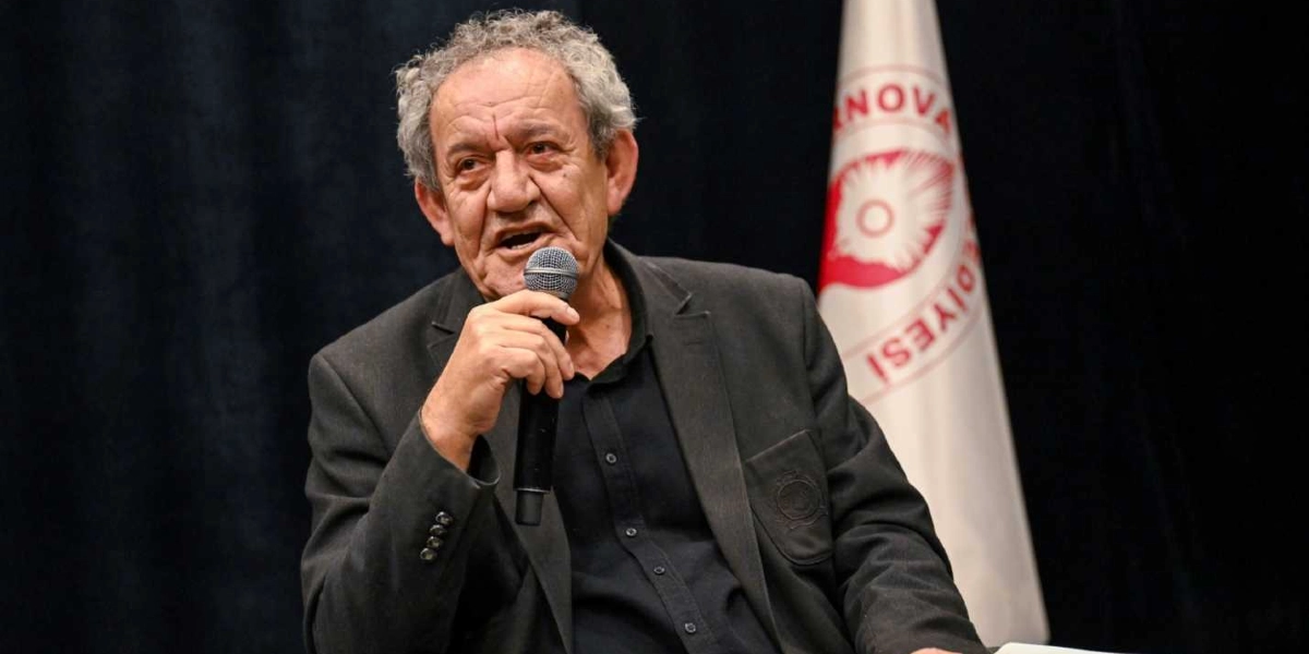 Koyu renkli bir ceket ve siyah gömlek giymiş, gri saçlı, orta yaşın üzerinde bir erkek, elinde mikrofon tutarak konuşma yapıyor. Arka planda siyah bir perde ve üzerinde Bornova Belediyesi logosunun bulunduğu beyaz bir bayrak yer alıyor.