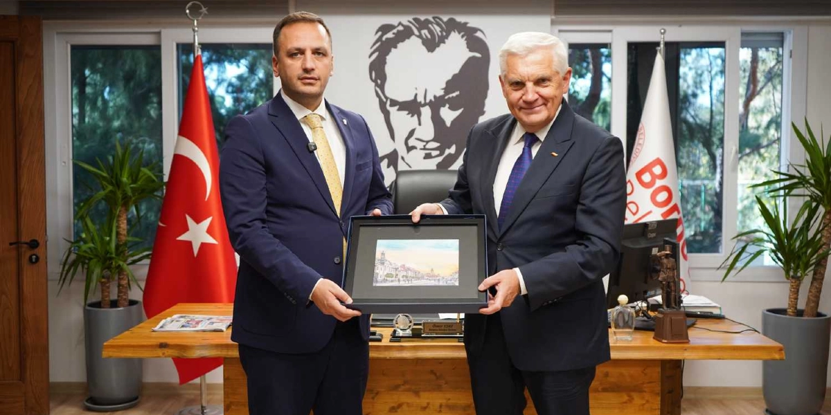 Takım elbiseli iki erkek, bir makam odasında yan yana durarak ellerinde çerçeveli bir tablo tutuyor. Arka planda Mustafa Kemal Atatürk'ün silüeti, Türk bayrağı ve Bornova Belediyesi'nin flaması yer alıyor. Masanın üzerinde küçük bir Atatürk heykeli ve isimlik bulunuyor.