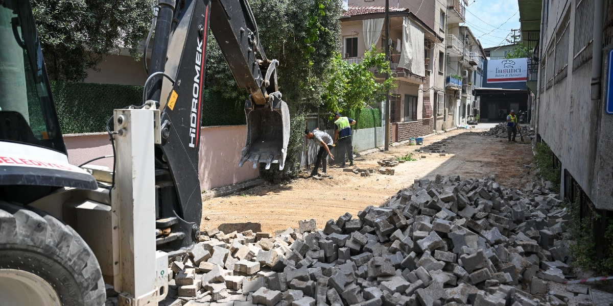 Çamdibi'nde bir sokakta belediyeye ait bir iş makinesi yol yenileme çalışması yapıyor. Ön planda sökülmüş parke taşlarından oluşan büyük bir yığın, arka planda ise işçilerin toprak zeminde çalıştığı bir sokak ve çevresindeki binalar görülüyor.