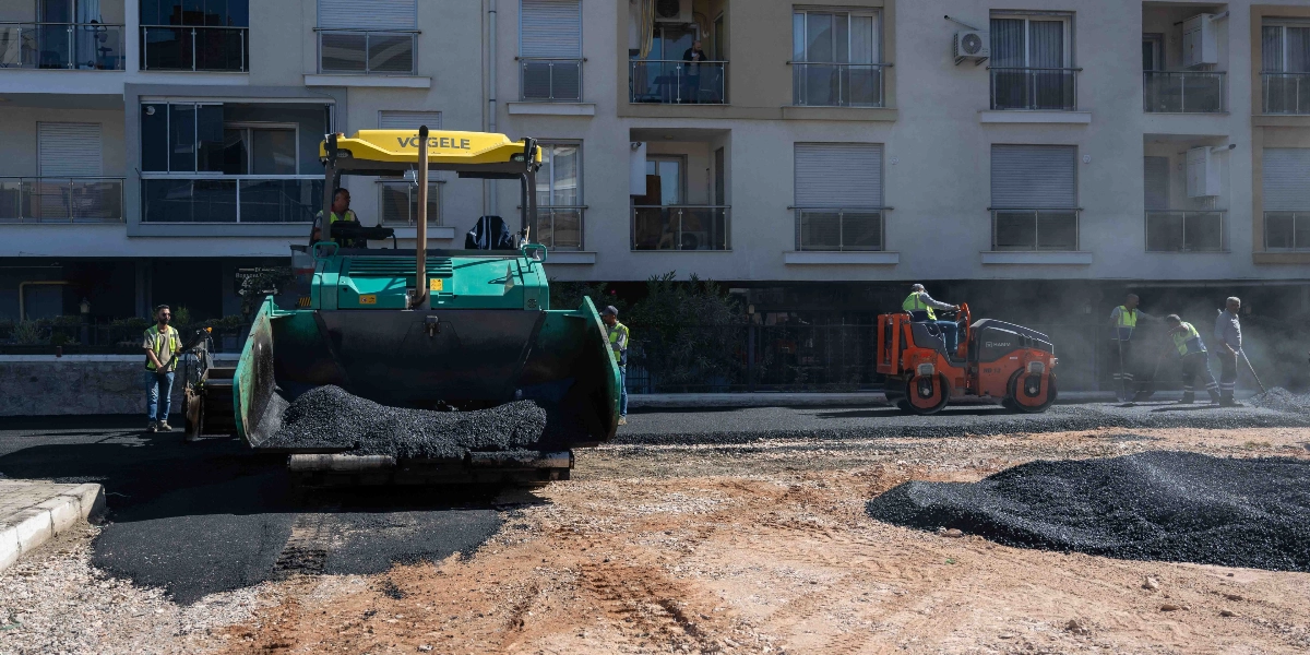 Erzene mahallesindeki bir sokakta, yeşil renkli bir Vögele asfalt serici makinesi ve turuncu bir silindir ile yol yenileme çalışması yapılıyor. İş makinelerinin başında ve çevresinde yelekli belediye çalışanları bulunuyor, zemin üzerine dökülen sıcak asfalt serilerek yol düzeltiliyor. Arka planda ise çok katlı bir apartman yer alıyor.