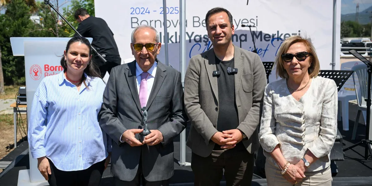 Bornova Belediyesi Sağlıklı Yaş Alma Merkezi etkinliğinde yan yana duran dört kişi. Soldan sağa; açık mavi çizgili gömlekli bir kadın, elinde küçük bir heykel tutan gri takım elbiseli ve sarı camlı gözlüklü bir beyefendi, gri ceketli bir erkek ve güneş gözlüklü, bej ceketli bir kadın yer alıyor. Arka planda Bornova Belediyesi logosunun bulunduğu bir kürsü ve 2024-2025 dönemine ait yazıların yer aldığı bir etkinlik panosu bulunuyor.