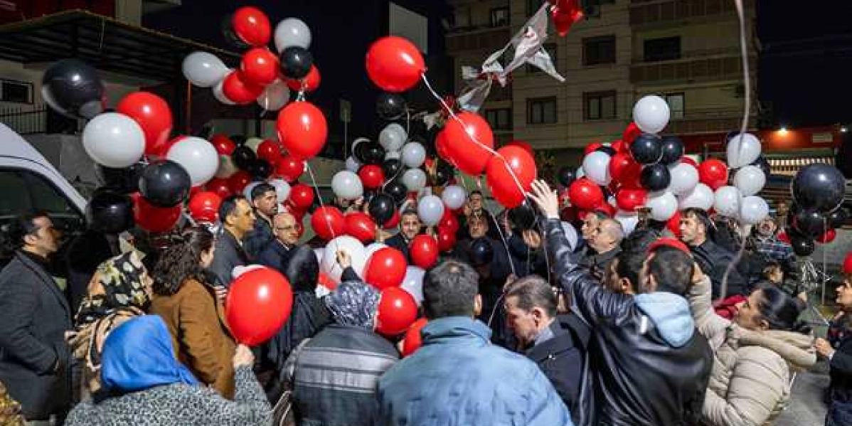 Gece vakti dış mekanda toplanmış bir grup insan, ellerinde tuttukları kırmızı, beyaz ve siyah renkli balonları gökyüzüne bırakıyor. Katılımcıların bir kısmı ellerindeki balonların iplerini havaya doğru uzatırken, bir kısmı da gökyüzüne yükselen balonları izliyor. Arka planda binaların görüldüğü anma etkinliğinde, insanlar yas ve anma atmosferini yansıtan bir topluluk oluşturuyor.