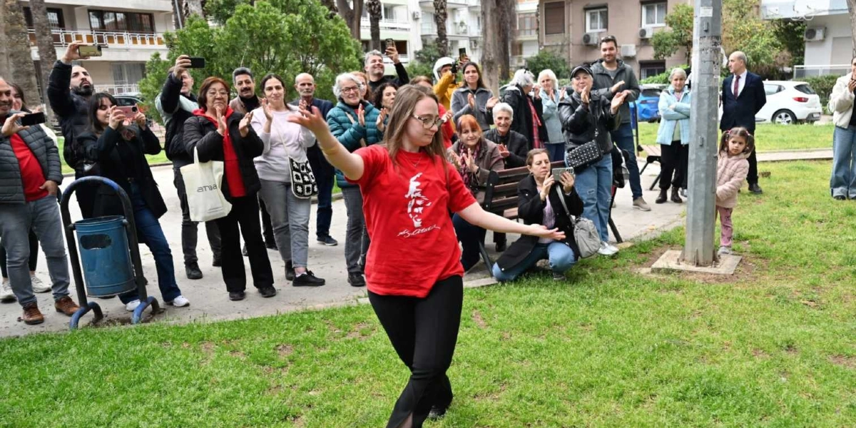 Açık havada, park benzeri bir alanda, üzerinde Atatürk silüeti bulunan kırmızı tişörtlü genç bir kadın dans figürü sergiliyor. Kadının etrafında toplanmış, çoğu alkışlayan ve cep telefonlarıyla çekim yapan kalabalık bir grup insan bulunuyor. Arka planda ağaçlar, banklar ve binalar yer alırken, sağ tarafta bir direğin arkasına saklanmış küçük bir kız çocuğu dikkat çekiyor.