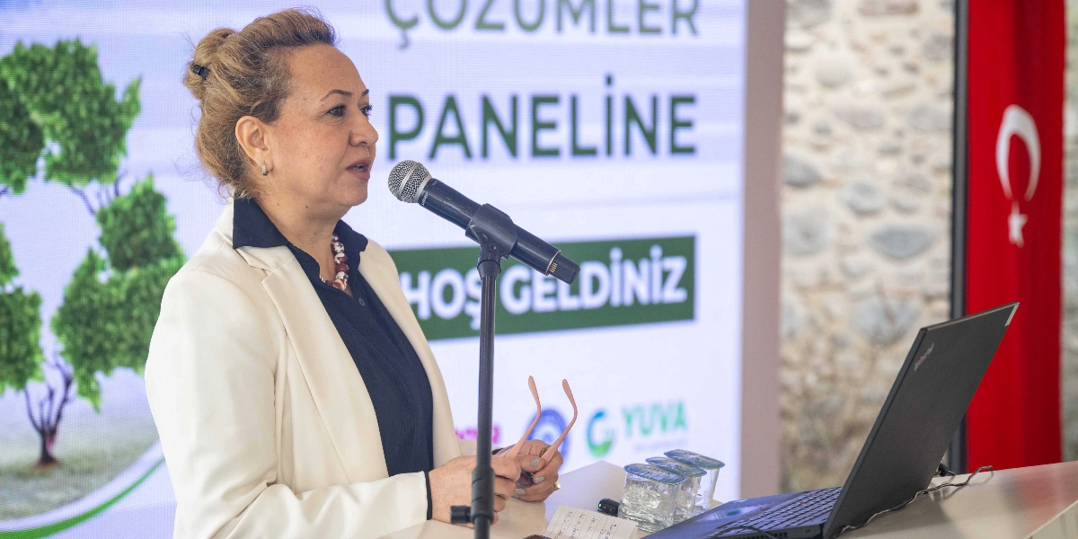 Krem rengi ceket ve koyu renkli bluz giymiş, saçlarını arkadan toplamış bir kadın, kürsüde mikrofon başında konuşma yapıyor. Kadının önünde açık bir dizüstü bilgisayar, bir not kağıdı ve su bardakları bulunuyor. Arka planda üzerinde 