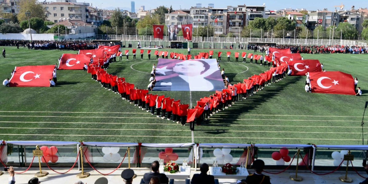 Bornova'da bir stadyumda düzenlenen Cumhuriyet Bayramı kutlamalarında, yeşil saha üzerinde toplanan kalabalık bir grup öğrenci, ellerindeki Türk bayraklarıyla büyük bir kalp şekli oluşturuyor. Kalbin merkezinde dev bir Mustafa Kemal Atatürk posteri yer alırken, çevresinde sıralanan öğrenciler ellerindeki bayrakları havaya kaldırarak görsel bir şölen sunuyor. Stadyumun tribünlerinde izleyiciler yer alırken, ön planda protokol üyeleri töreni takip ediyor.