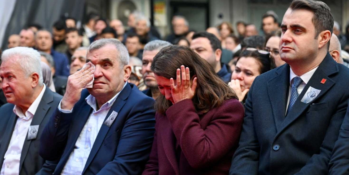 Kalabalık bir topluluk önünde, ceketlerinin yakasında anma fotoğrafı bulunan kişiler duygusal anlar yaşıyor. Ön sırada oturan gri saçlı bir adam elindeki mendille gözyaşlarını silerken, yanındaki bordo ceketli kadın yüzünü elleriyle kapatarak ağlıyor. En sağda ise koyu renk takım elbiseli bir erkek, hüzünlü bir ifadeyle karşıya bakıyor.