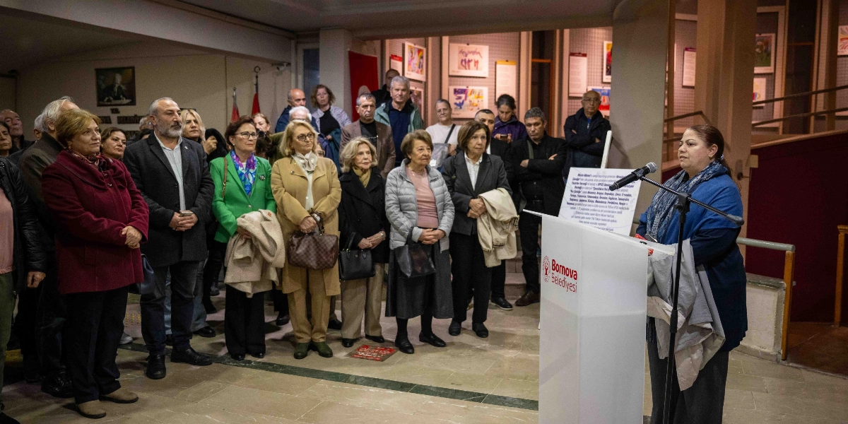 Bornova Belediyesi'nin düzenlediği bir etkinlikte, kürsüde konuşma yapan bir kadın ve onu dinleyen katılımcı grubu. Kürsünün üzerinde Bornova Belediyesi logosu ve Nazım Hikmet ile ilgili bilgilendirici bir metin yer alıyor. Arka planda ise duvarlarda sergilenen sanat eserleri ve izleyiciler bulunuyor.