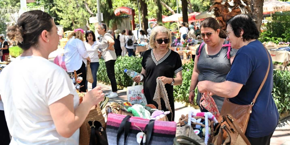 Açık havada kurulan bir el sanatları pazarında, el yapımı örgü çantalar ve ürünlerin sergilendiği bir tezgahın önünde duran üç kadın, ürünleri inceliyor. Ön planda, beyaz tişörtlü bir kadın, elindeki hasır çantayı müşterilere gösterirken, güneş gözlüklü ve siyah elbiseli bir kadın elinde tuttuğu örgü bir parçayı inceliyor. Yanındaki diğer kadın ise ürünleri dikkatle izliyor. Arka planda ise ağaçlar ve pazar alanında dolaşan diğer insanlar görülüyor.