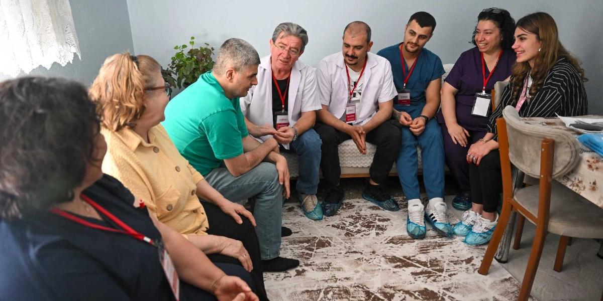 Ev ortamında bir araya gelmiş, aralarında sağlık personeli olduğu anlaşılan bir grup insan sohbet ediyor. Sağ taraftaki koltukta oturan beyaz önlüklü iki kişi, mavi üniformalı bir erkek, mor formasıyla bir kadın ve çizgili ceketli genç bir kadın, yeşil tişörtlü bir adam ve sarı hırkalı bir kadınla karşılıklı oturarak iletişim kuruyor. Bazı kişilerin boyunlarında kimlik kartları asılı ve ayaklarında galoş bulunuyor. Ortamda samimi ve destekleyici bir atmosfer hakim.