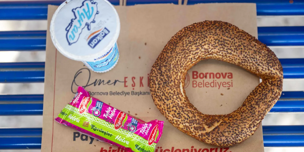 Mavi bir bankın üzerine yerleştirilmiş Bornova Belediyesi logolu kese kağıdının üzerinde bir adet susamlı simit, bir paket Kardeşler tahin helvası ve bir kutu Balkan ayran bulunuyor.