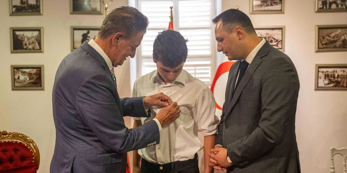 Gri takım elbiseli yaşlı bir adam, beyaz gömlekli genç bir çocuğun göğsüne rozet takıyor. Yanlarında koyu renk takım elbiseli ve kravatlı bir başka adam, ellerini önünde birleştirmiş şekilde bu anı izliyor. Arka planda duvarlarda siyah beyaz tarihi fotoğraflar asılı ve kısmen görünen bir Türk bayrağı bulunuyor.