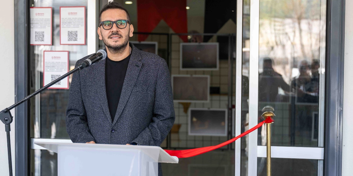 Siyah tişört ve üzerine gri desenli bir ceket giymiş, gözlüklü bir erkek, bir binanın girişinde kürsüde konuşma yapıyor. Kürsünün önünde açılış törenlerini simgeleyen kırmızı bir kurdele şeridi bulunuyor. Arka planda cam kapıların üzerinde Bornova Belediyesi logolu bilgilendirme panoları ve içeride sergilenen fotoğraflar yer alıyor.