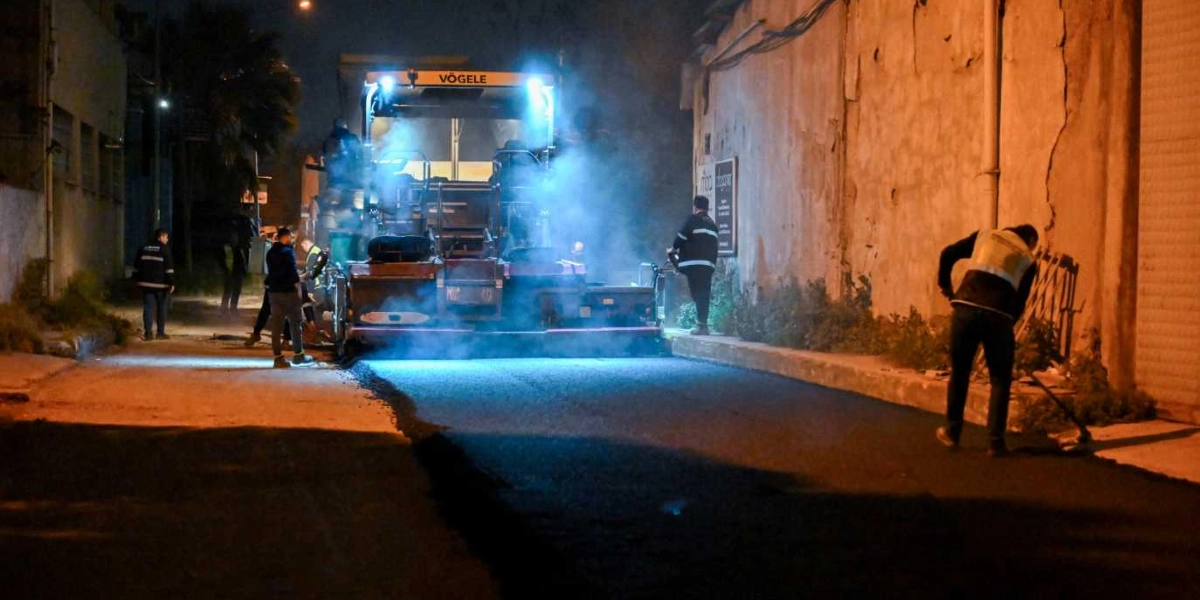Gece vakti dar bir sokakta asfalt serme çalışması yapan Vögele marka bir finişer makinesi ve çevresinde çalışan işçiler görülüyor. Makineden yükselen dumanlar ışık altında belirginleşirken, işçilerden biri sağ tarafta asfaltı düzeltmek için elindeki aletle çalışıyor.
