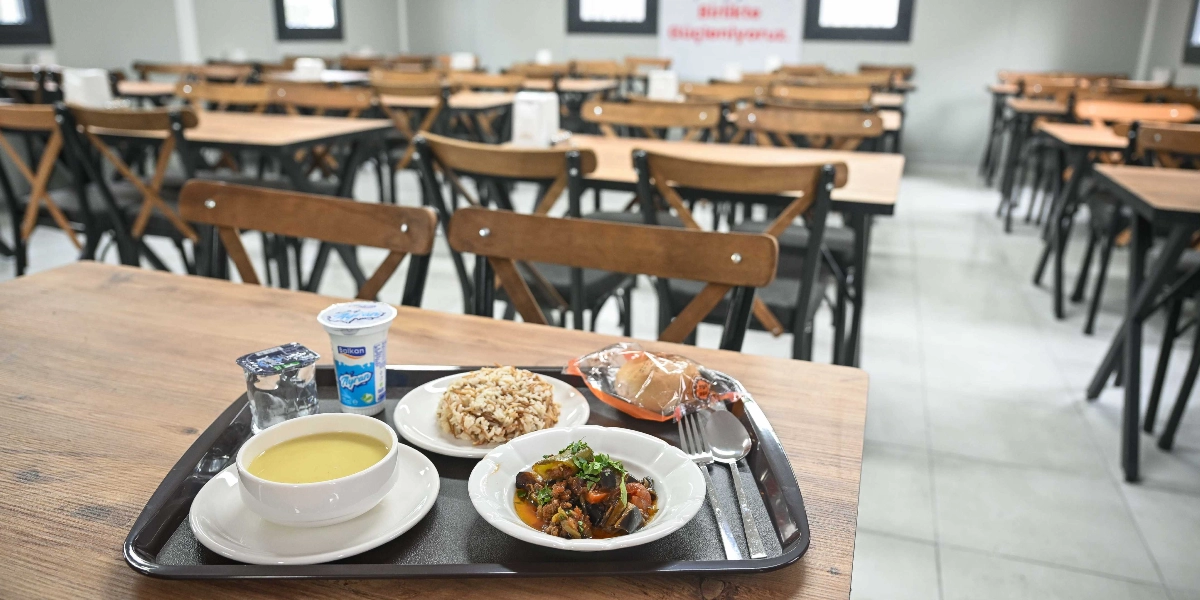 Modern ve geniş bir yemekhanede, ön plandaki ahşap masanın üzerinde duran siyah bir tepsi içerisinde servis edilmiş öğle yemeği. Tepside bir kase çorba, bir tabak pilav, bir tabak kıymalı patlıcan yemeği, bir paket ekmek, bir bardak su ve bir kutu ayran bulunuyor. Arka planda ise düzenli bir şekilde dizilmiş çok sayıda boş ahşap masa ve sandalye yer alıyor.
