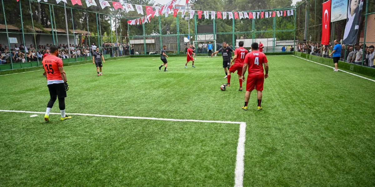 Açık havada, etrafı tellerle çevrili ve Türk bayrakları ile süslenmiş bir halı sahada futbol maçı oynanıyor. Kırmızı formalar giyen oyuncular sahada top sürme ve paslaşma halindeyken, kaleci Sinan 35 numaralı formasıyla kalesinde bekliyor. Saha kenarındaki teller arkasında maçı izleyen seyirciler bulunuyor.