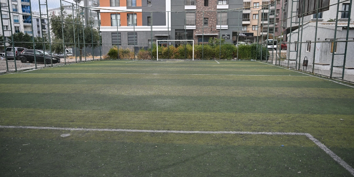 Etrafı yüksek tellerle çevrili, suni çim zeminli, içerisinde beyaz bir futbol kalesi bulunan boş bir halı saha. Saha, yerleşim yerlerinin ve apartmanların arasında yer alıyor.