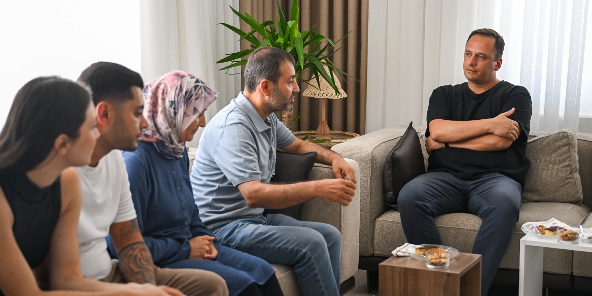 Aydınlık bir oturma odasında, bej renkli koltuklarda yan yana oturan dört kişilik bir grup ve karşısında tek başına oturan bir adam sohbet ediyor. Soldan sağa doğru, siyah kolsuz bir bluz giymiş genç bir kadın, beyaz tişörtlü dövmeli bir genç erkek, başörtülü orta yaşlı bir kadın ve açık mavi polo yaka tişörtlü, sakallı bir adam bir arada oturuyor. Karşılarındaki koltukta ise siyah tişörtlü ve lacivert pantolonlu bir adam, kollarını kavuşturmuş, ciddi bir ifadeyle onları dinliyor. Önlerindeki küçük sehpalarda ikramlıklar bulunuyor.