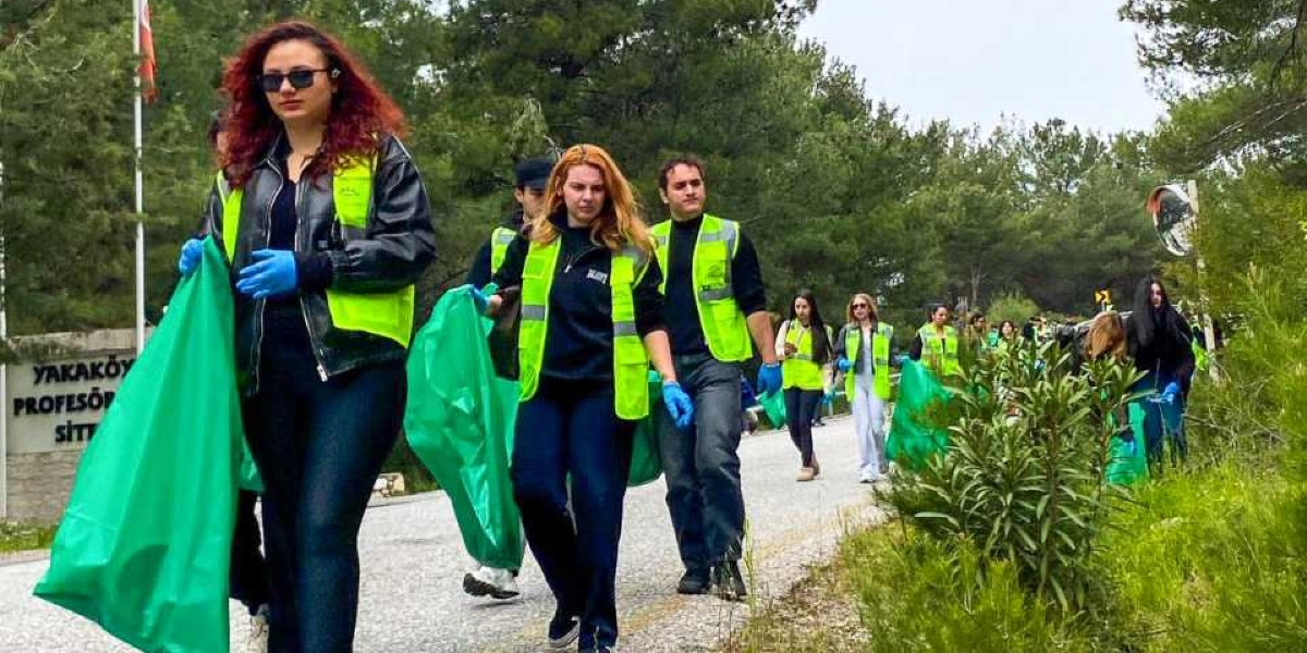 Üzerlerinde neon sarı yelekler ve ellerinde mavi eldivenler bulunan bir grup gönüllü, ellerindeki büyük yeşil çöp poşetleriyle ormanlık bir yol kenarında temizlik çalışması yapıyor. Arka planda 