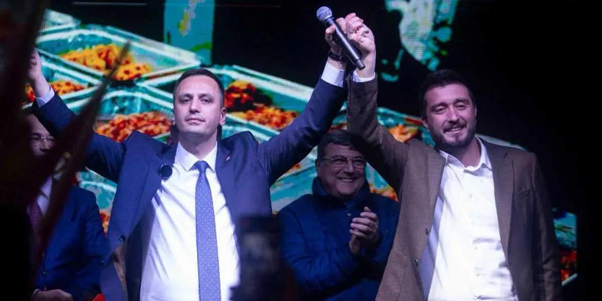 Lacivert takım elbiseli ve kravatlı bir adam ile kahverengi ceketli bir adam, sahnede yan yana durarak ellerini havaya kaldırmış selamlıyor. Sağ taraftaki adam elinde bir mikrofon tutuyor. Arka planda büyük bir dijital ekran ve onları izleyen, alkışlayan bir kişi daha bulunuyor.