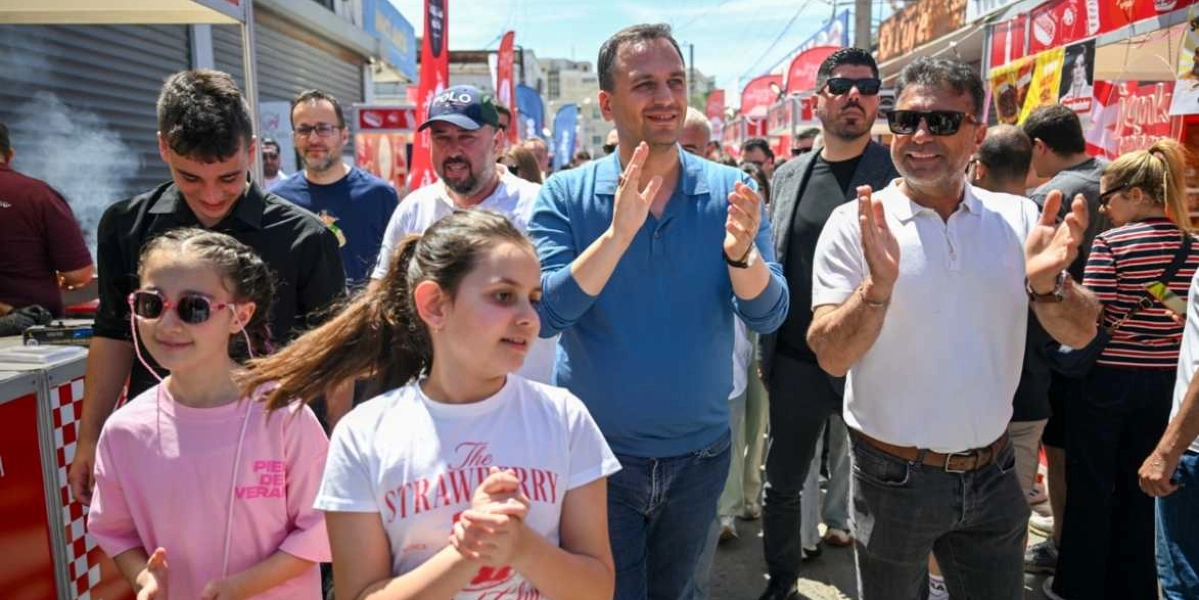 Bornova'daki bir festival alanında, aralarında Bornova Belediye Başkanı Ömer Eşki'nin de bulunduğu bir grup insan, yürüyüş yaparken alkışlayarak etkinliğe katılıyor. Ön planda pembe ve beyaz tişörtlü iki kız çocuğu, arka planda ise çeşitli stantların ve kalabalığın olduğu güneşli bir açık hava ortamı görülüyor.