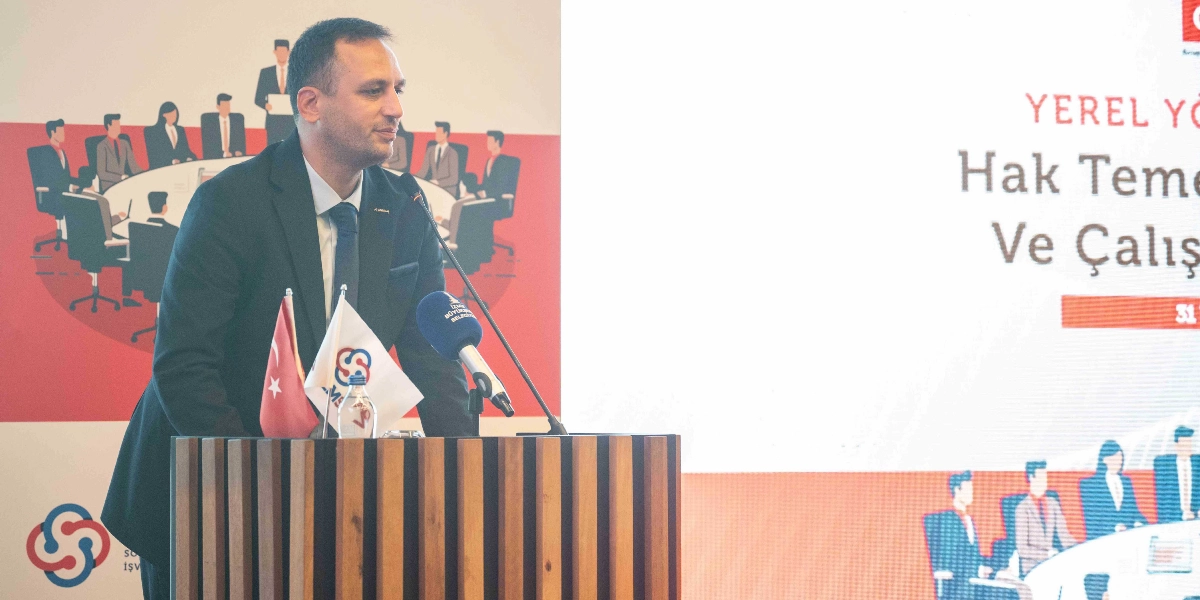 İzmir Büyükşehir Belediye Başkanı Cemil Tugay, SODEMSEN tarafından düzenlenen çalıştayda kürsüde konuşma yapıyor. Önünde Türk bayrağı ve SODEMSEN logolu bir masa bayrağı bulunan Başkan Tugay'ın arkasındaki ekranda 