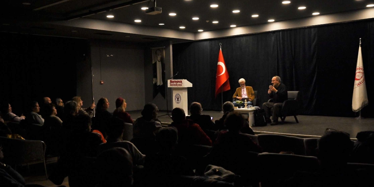 Bornova Belediyesi tarafından düzenlenen bir söyleşi etkinliğinde, sahnedeki iki konuşmacı koltuklarda oturarak izleyicilere hitap ediyor. Sahnenin ortasında üzerinde Bornova Belediyesi logosu bulunan bir kürsü, arkasında ise Türk bayrağı ve Bornova Belediyesi flaması yer alıyor. Arka duvarda Atatürk'ün bir portresi asılıyken, loş ışıklı salonda izleyiciler sıralar halinde oturarak konuşmacıları dinliyor.
