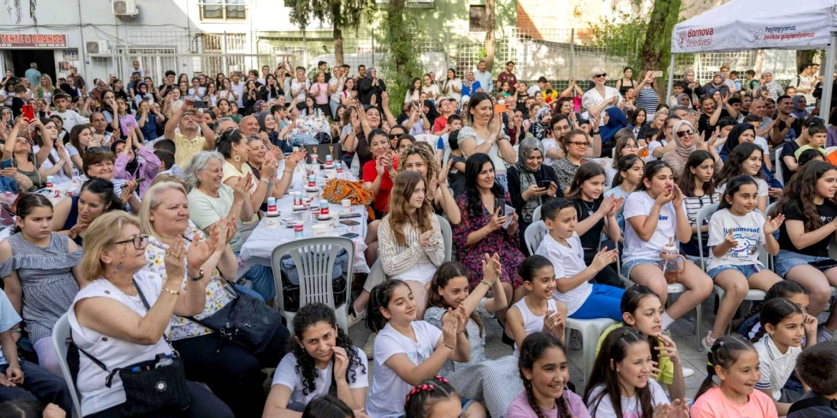Açık havada düzenlenen bir etkinlikte, sandalyelerde oturan ve büyük bir kısmı kadınlar ile çocuklardan oluşan kalabalık bir grup, alkışlayarak bir gösteriyi izliyor. İnsanlar masaların etrafına dizilmiş, neşeli bir atmosferde bir araya gelmiş durumda. Arka planda Bornova Belediyesi'ne ait bir çadır ve ağaçlar yer alıyor.