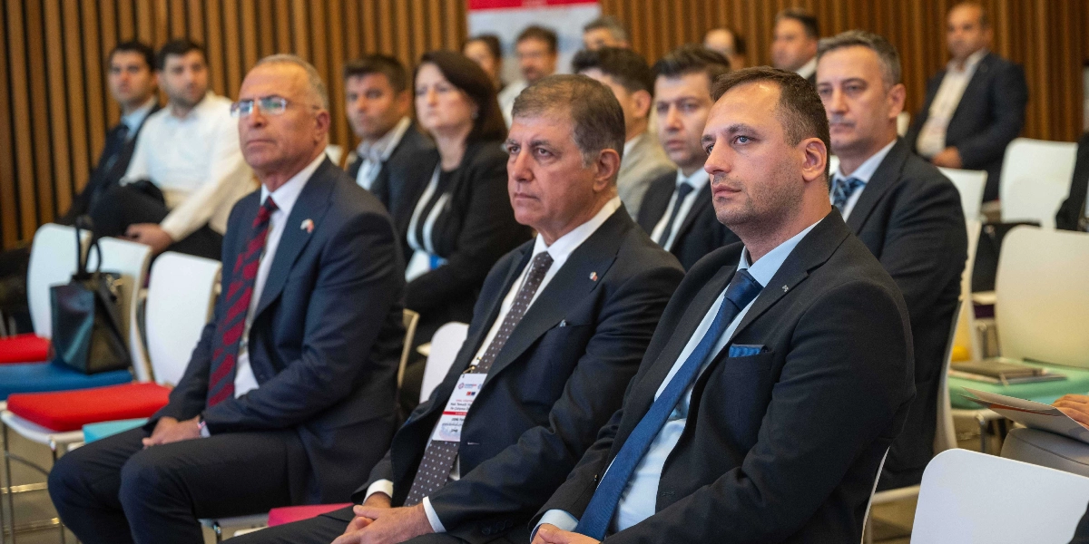 SODEMSEN çalıştayında, takım elbiseli katılımcılar bir salonda oturmuş dikkatle konuşmacıyı dinliyor. Ön planda, koyu renk takım elbiseli ve lacivert kravatlı üç erkek yan yana oturuyor; aralarında Bornova Belediye Başkanı Ömer Eşki de yer alıyor. Arka sıralarda ise farklı katılımcılar, ahşap panelli bir duvarın önünde, beyaz sandalyelerde oturarak etkinliği takip ediyor.