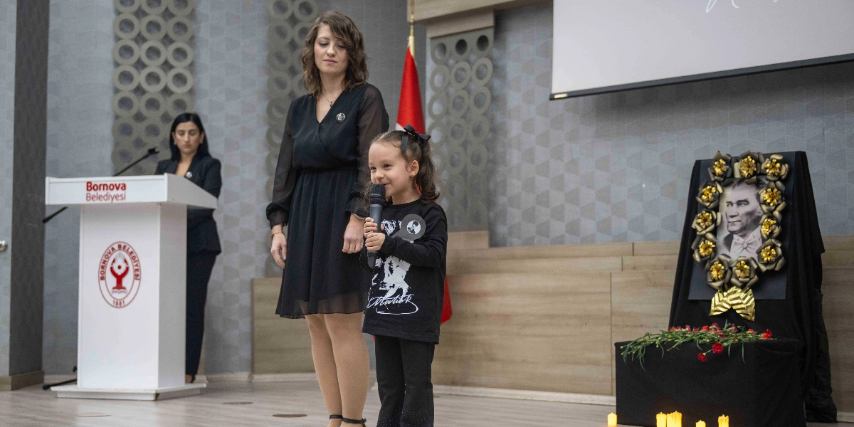 Bornova Belediyesi'ne ait bir salonda düzenlenen anma töreninde, üzerinde Atatürk baskılı siyah bir tişört olan küçük bir kız çocuğu elinde mikrofonla konuşma yapıyor. Çocuğun yanında siyah elbiseli bir kadın duruyor. Arka planda Bornova Belediyesi logosunun bulunduğu bir kürsü, Türk bayrağı ve sağ tarafta siyah çiçeklerle çevrili bir Atatürk portresi ile önünde kırmızı karanfiller ve mumlar yer alıyor.