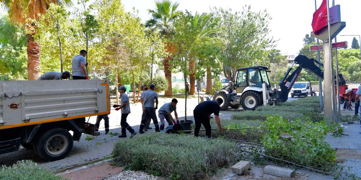Bornova Belediyesi çalışanları, park alanında yeni fidan dikimi ve çevre düzenlemesi yapıyor. İşçiler, kamyondan indirdikleri genç ağaçları saksılarından çıkararak toprağa yerleştiriyor. Çalışma alanında bir iş makinesi ve arka planda ağaçlıklı bir park görünümü yer alıyor. Sağ taraftaki direğin tepesinde dalgalanan bir Türk bayrağı bulunuyor.