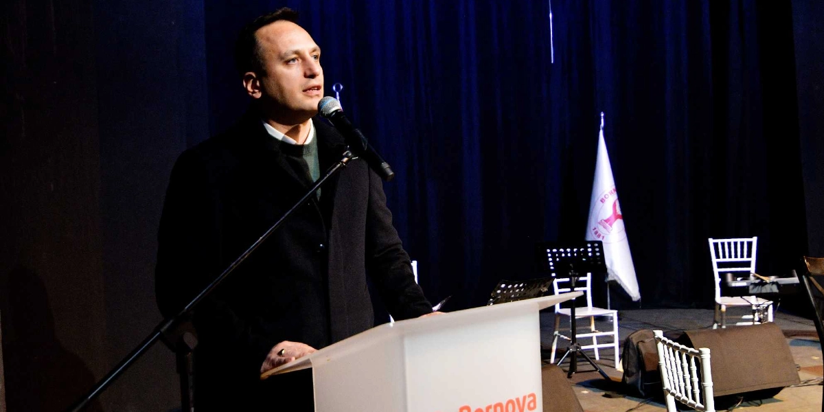 Koyu renkli bir ceket giymiş bir erkek, üzerinde Bornova Belediyesi logosu bulunan beyaz bir kürsüde mikrofon başında konuşma yapıyor. Arka planda lacivert perdeler, beyaz bir bayrak ve sahnede yer alan beyaz sandalyeler görülüyor.