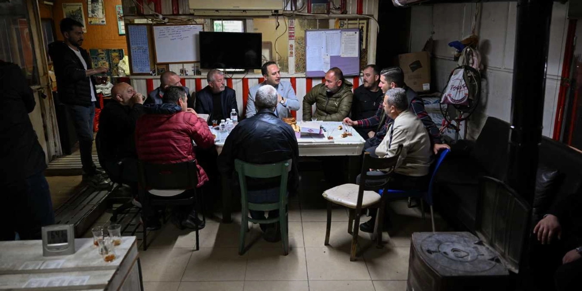 Loş ışıklı bir mekanda, beyaz bir masa etrafında toplanmış yaklaşık on kişilik bir erkek grubu sohbet ediyor. Masanın üzerinde çay bardakları ve yiyecekler bulunuyor. Arka planda duvarda asılı bir televizyon, çeşitli notlar ve ilanlar yer alıyor. Grubun bir kısmı otururken, biri ayakta durarak onlara eşlik ediyor. Ortam, mütevazı bir esnaf lokantası veya dernek lokali havası taşıyor.