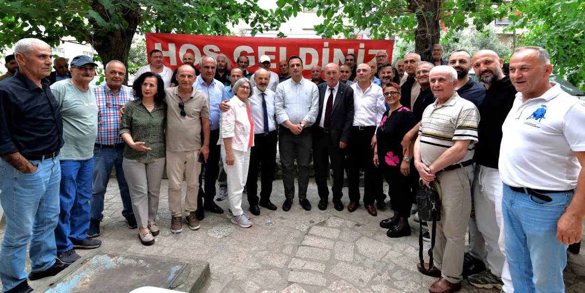 Açık havada, ağaçların altında bir araya gelmiş kalabalık bir grup insan, arkalarında üzerinde beyaz harflerle 