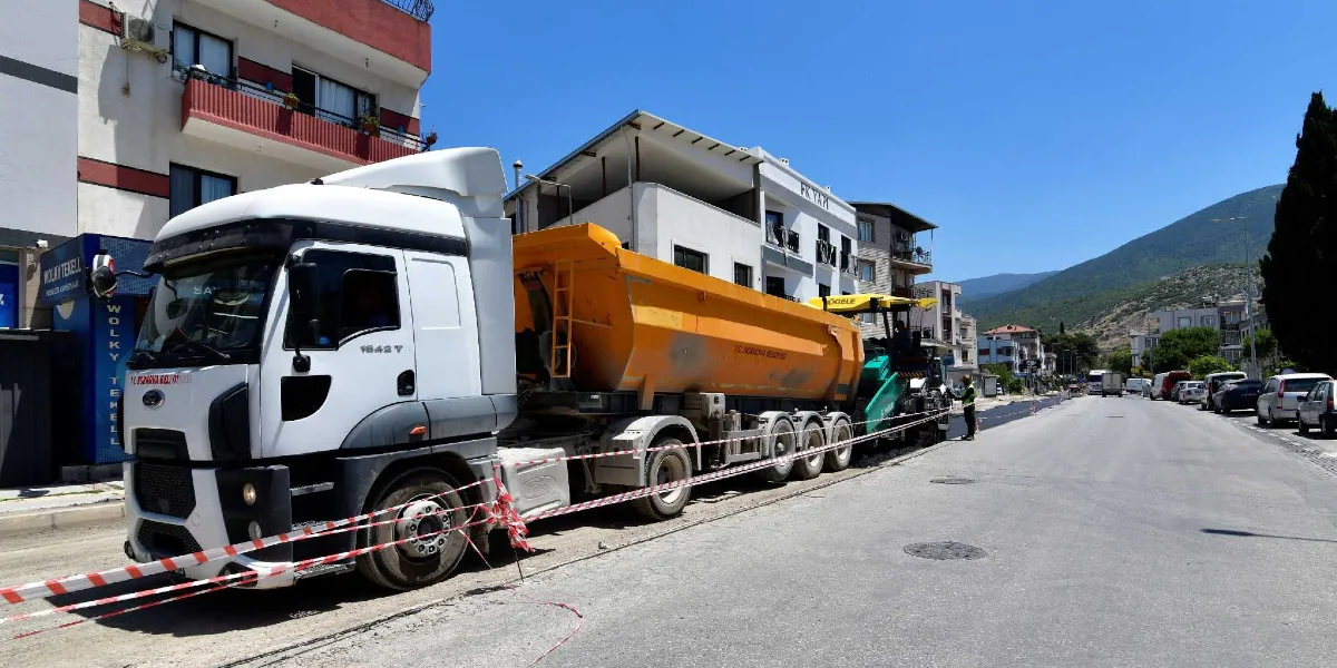 Bornova Belediyesi'ne ait beyaz bir çekici ve ona bağlı sarı bir damperli kamyon, yol kenarında park halinde duruyor. Kamyonun arkasında, asfalt serme çalışması yapan yeşil renkli bir finişer makinesi ve başında çalışan bir görevli bulunuyor. Çalışma alanı, kırmızı beyaz şeritlerle çevrilerek güvenlik altına alınmış durumda. Arka planda ise apartmanlar, ağaçlar ve dağ manzarası görülüyor.