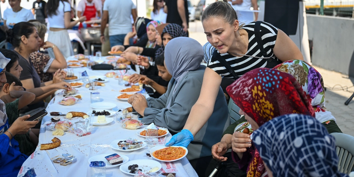 Açık havada uzun bir masada toplanmış bir grup insan, birlikte kahvaltı yapıyor. Siyah beyaz çizgili tişört giyen ve elinde mavi eldiven bulunan bir kadın, masadaki kişilere tabakla yemek ikram ediyor. Masanın üzerinde simit, zeytin, peynir ve menemen gibi çeşitli kahvaltılıklar bulunuyor. Katılımcıların birçoğu başörtülü kadınlardan ve çocuklardan oluşuyor.