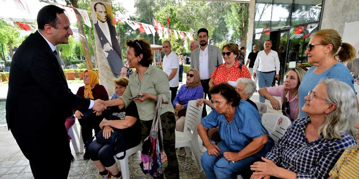 Siyah takım elbiseli bir erkek, açık havada düzenlenen bir etkinlikte, ayakta duran ve elinde telefon tutan bir kadınla tokalaşıyor. Etrafında beyaz plastik sandalyelerde oturan ve ayakta duran çeşitli yaş gruplarından kadınlar bulunuyor. Arka planda Mustafa Kemal Atatürk'ün büyük bir posteri ve Türk bayraklarıyla süslenmiş bir alan görülüyor.